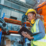 Female holding laptop while working next to robotic arm in advanced manufacturing facility.