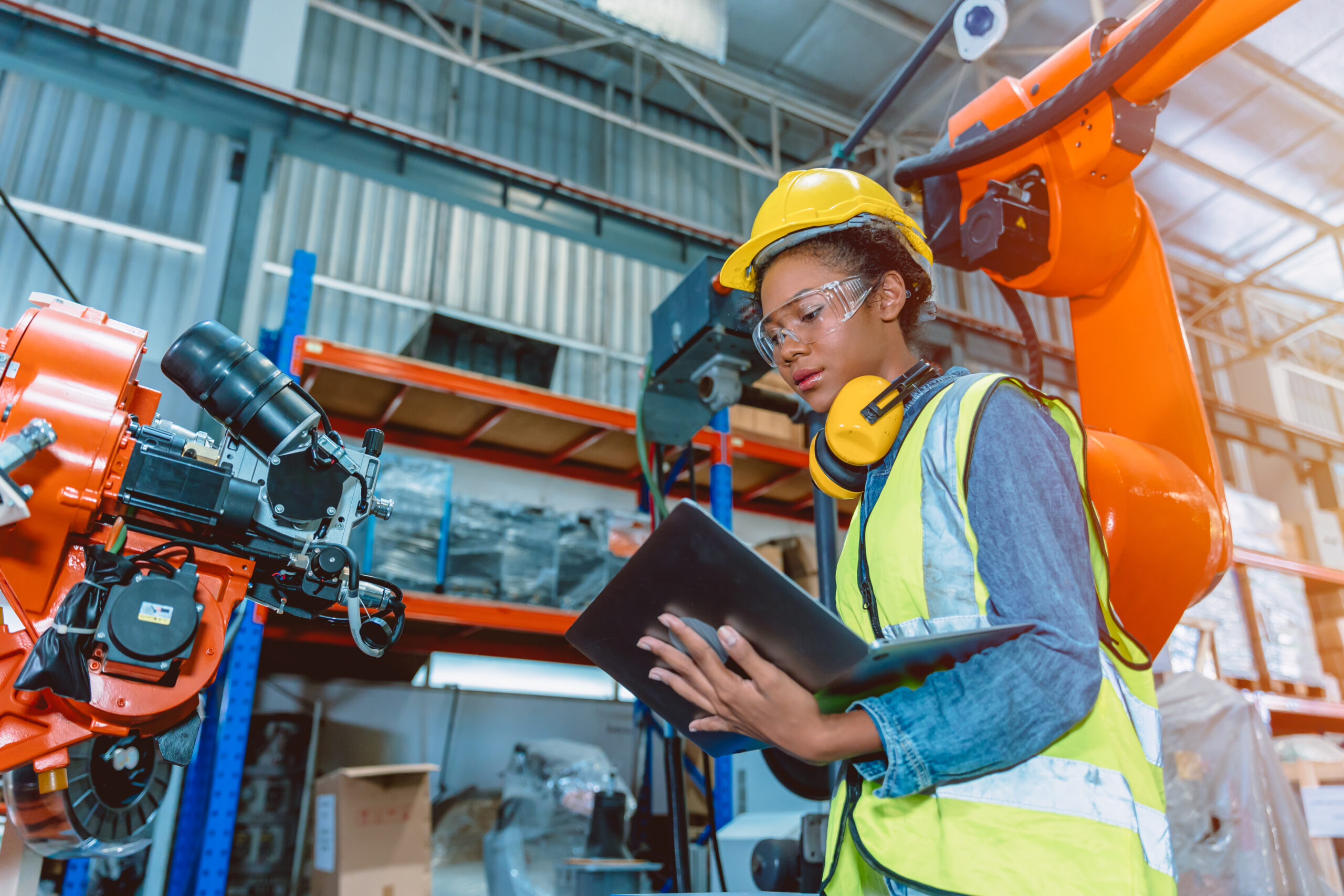 Female holding laptop while working next to robotic arm in advanced manufacturing facility.